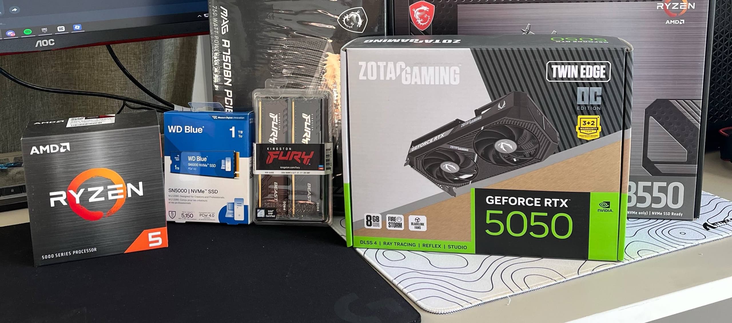 Boxed components on a desk: a Ryzen 5 processor, a WD Blue NVMe drive, Kingston Fury memory, a Zotac GeForce RTX 5050 and an MSI B550 motherboard.