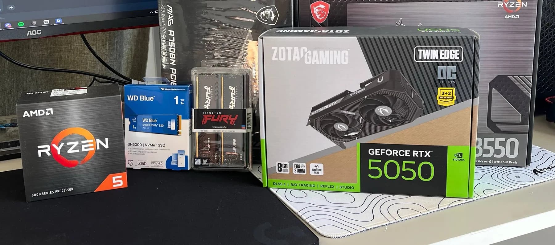 Boxed components on a desk: a Ryzen 5 processor, a WD Blue NVMe drive, Kingston Fury memory, a Zotac GeForce RTX 5050 and an MSI B550 motherboard.